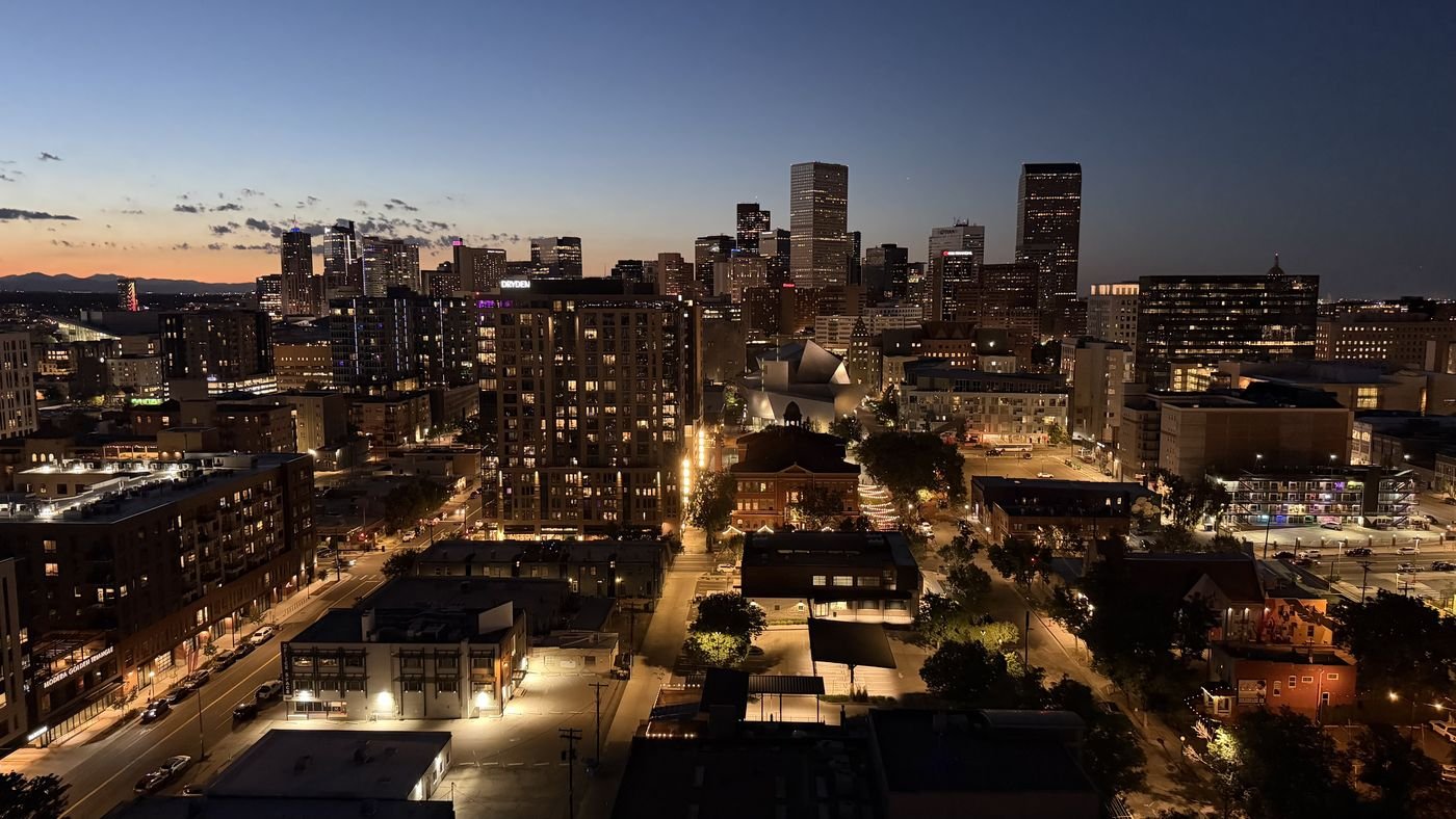 Downtown Denver at dusk from above: lit office towers in the middle distance, warm street lights in the low brick blocks in front of them, and the Front Range foothills silhouetted against an orange sunset on the horizon.